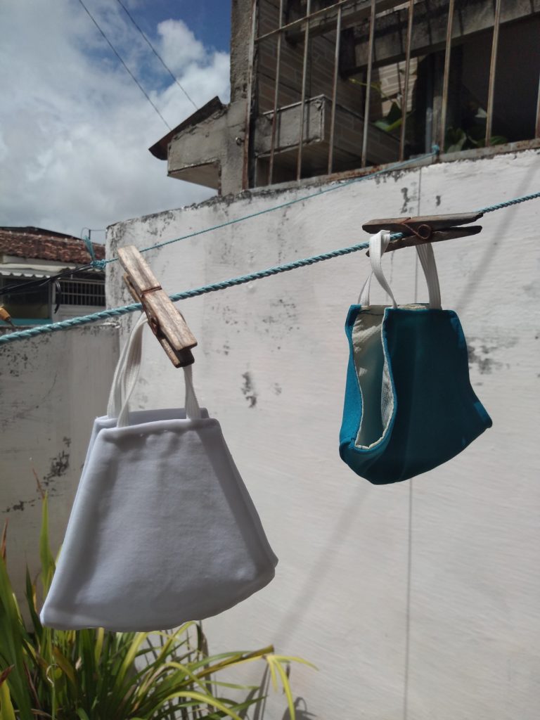 Máscaras 1 Imagem de duas máscaras de pano penduradas em um varal, uma branca e uma azul. Fim da descrição.