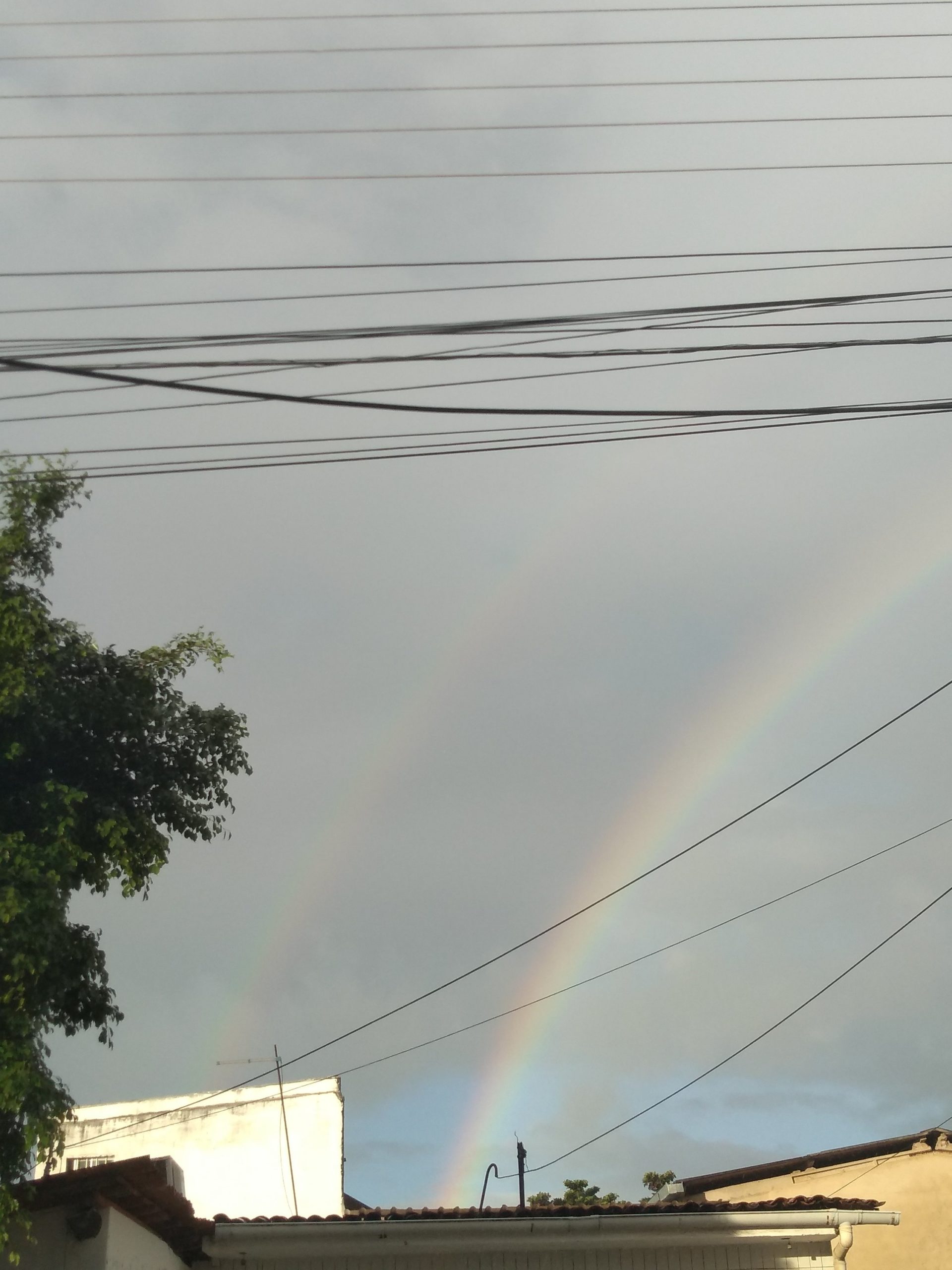 Vocês já viram um duplo arco-íris? 1 Foto do céu visto da rua, onde se veem dois arco-íris. Fim da descrição.