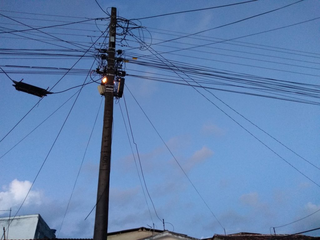 Nem todo anoitecer é igual na varanda 1 Imagem do céu ao anoitecer, poste com fios elétricos e de telefone com a luz acesa, ao fundo os telhados das casas da rua. Fim da descrição.