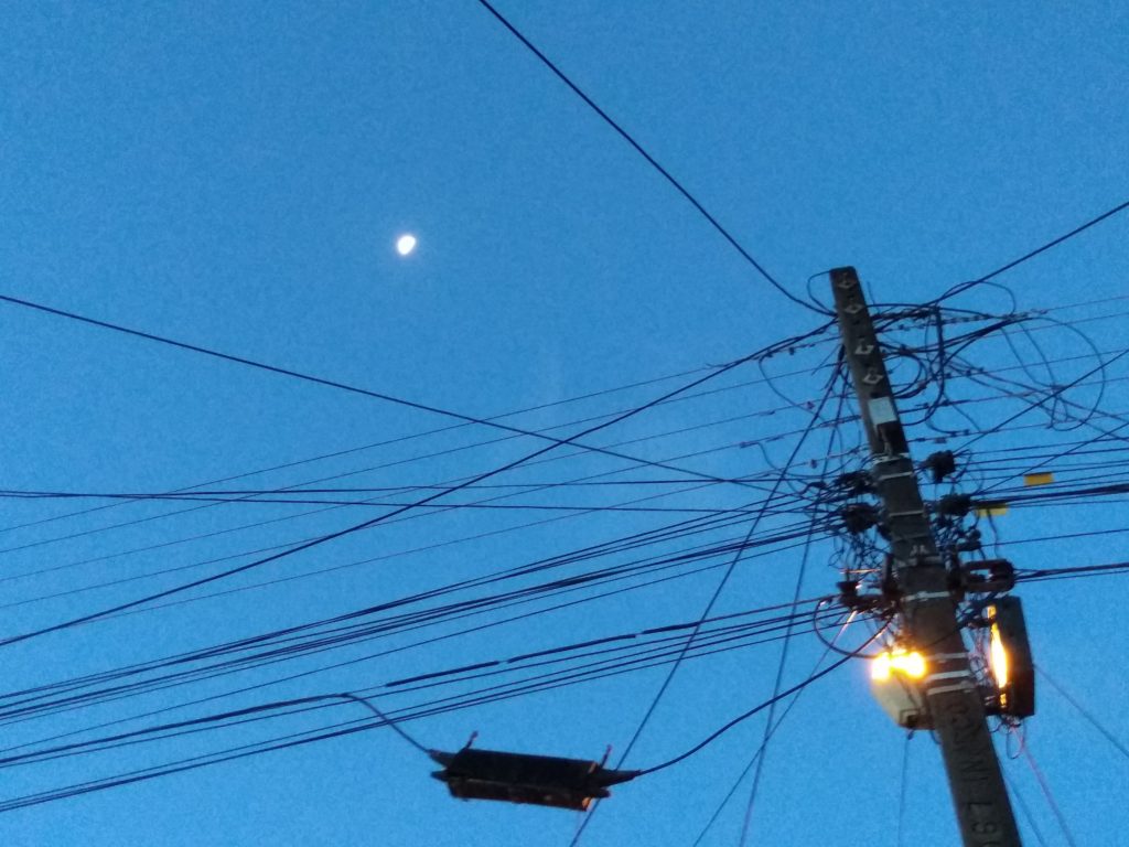 Nem todo anoitecer é igual na varanda 2 Imagem do céu na hora do anoitecer, com a Lua Crescente ao fundo e um poste com fios elétricos e de telefone com a luz acesa. Fim da descrição.